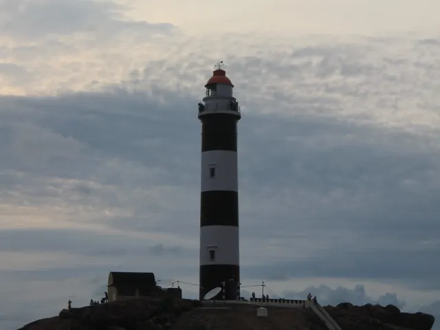 The striped Kapu lighthouse on its rock under a cloudy sky